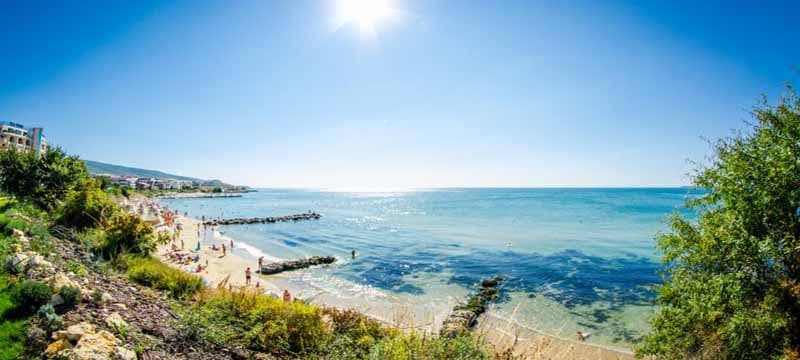 A bright sunny day on the Bulgarian Black Sea coast, with golden sand, turquoise water, and visitors enjoying the beach surrounded by lush greenery and clear blue skies.