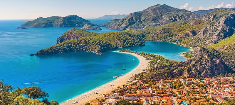 A breathtaking aerial view of the Ölüdeniz Blue Lagoon in Fethiye, Turkey, with turquoise waters, golden sandy beaches, lush green hills, and the dramatic mountains of the Turquoise Coast under a clear blue sky.