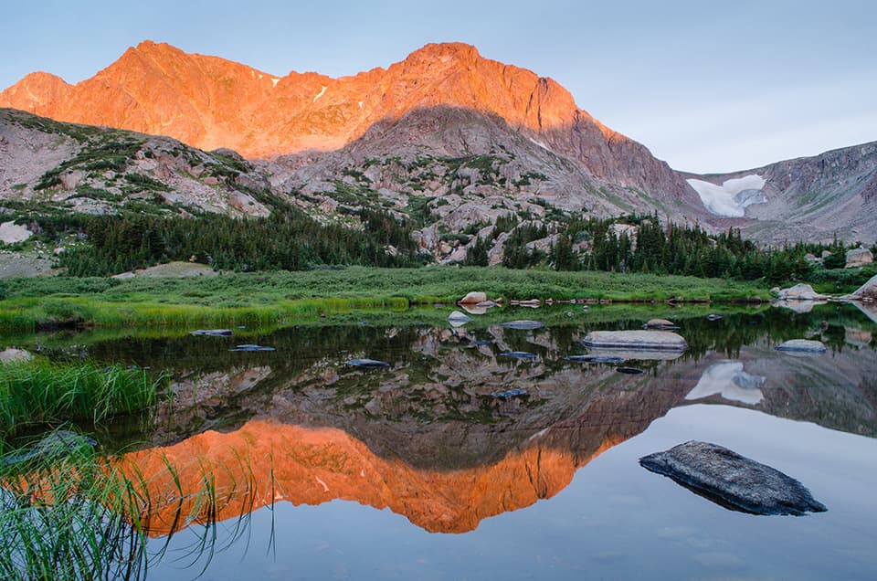 Sunrise over Rocky Mountain National Park showing a serene lake reflection with lush greenery and a towering, sunlit mountain backdrop, encapsulating Colorado's natural beauty.